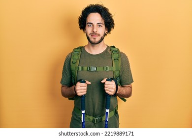 Handsome Hispanic Man Wearing Backpack Holding Trekking Poles Relaxed With Serious Expression On Face. Simple And Natural Looking At The Camera. 