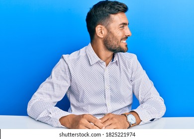 Handsome Hispanic Man Wearing Business Clothes Sitting On The Table Looking To Side, Relax Profile Pose With Natural Face And Confident Smile. 