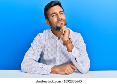 Handsome Hispanic Man Wearing Business Clothes Sitting On The Table Looking Confident At The Camera Smiling With Crossed Arms And Hand Raised On Chin. Thinking Positive. 