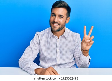 Handsome Hispanic Man Wearing Business Clothes Sitting On The Table Smiling Looking To The Camera Showing Fingers Doing Victory Sign. Number Two. 