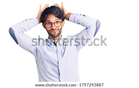 Handsome hispanic man wearing business shirt and glasses doing bunny ears gesture with hands palms looking cynical and skeptical. easter rabbit concept. 