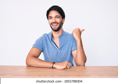 Handsome Hispanic Man Wearing Casual Clothes Sitting On The Table Smiling With Happy Face Looking And Pointing To The Side With Thumb Up. 