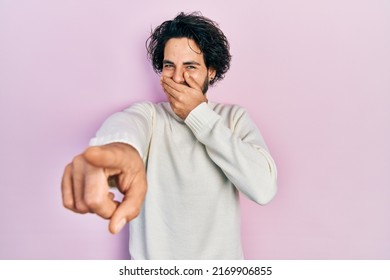 Handsome Hispanic Man Wearing Casual White Sweater Laughing At You, Pointing Finger To The Camera With Hand Over Mouth, Shame Expression 