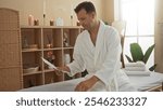 Handsome hispanic man in a white robe sitting in a spa room, reading a brochure surrounded by candles and plants