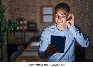 Handsome Hispanic Man Working At The Office At Night Smiling With Hand Over Ear Listening An Hearing To Rumor Or Gossip. Deafness Concept. 