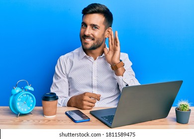 Handsome Hispanic Man Working Using Laptop At The Office Smiling With Hand Over Ear Listening And Hearing To Rumor Or Gossip. Deafness Concept. 