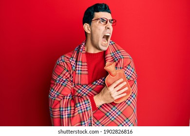 Handsome Hispanic Man Wrapped In A Winter Blanket Holding Hot Water Bag Angry And Mad Screaming Frustrated And Furious, Shouting With Anger Looking Up. 