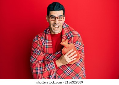 Handsome hispanic man wrapped in a winter blanket holding hot water bag smiling and laughing hard out loud because funny crazy joke. 