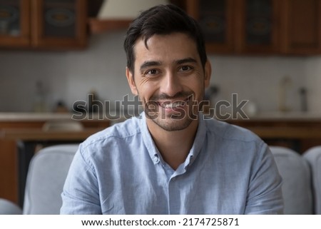 Handsome Hispanic millennial man sit on sofa at home look at camera. Make video call, remote talk, profile picture of video conference streamer, homeowner portrait, tenancy, modern flat owner concept