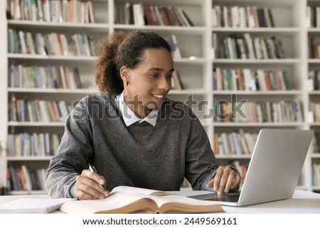 Handsome Hispanic student guy e-learning, using laptop, takes notes in copybook, prepare assignment or essay in library, smile, enjoy studying process. Generation Z and education, modern technology