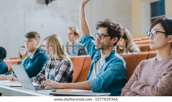 Handsome Hispanic\
Student Uses Laptop while Listening to a Lecture at the University,\
He Raises Hand and Asks Lecturer a Question. Multi Ethnic Group of\
Modern Bright\
Students.