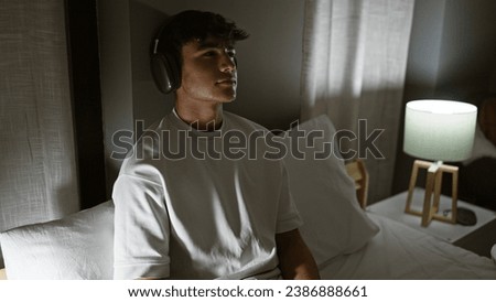 Handsome hispanic teenager enjoying his favorite song, relaxed in his cozy bedroom, lounging on his comfortable bed at night with the dim light of the lamp