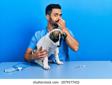 Handsome hispanic veterinary man with beard checking dog health smelling something stinky and disgusting, intolerable smell, holding breath with fingers on nose. bad smell 