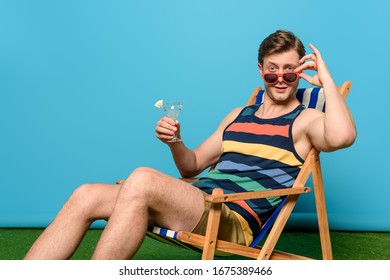 handsome holding glass of cocktail, touching glasses and looking at camera while sitting in deck chair on blue background - Shutterstock ID 1675389466