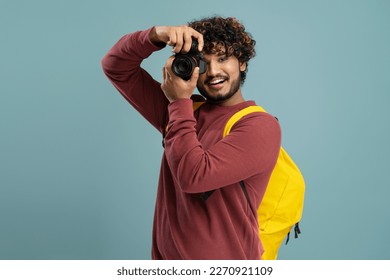 Handsome Indian curly-haired young man with yellow backpack, a student photographer using his modern digital camera and smiling cheerfully while taking photo, isolated over blue color background - Powered by Shutterstock