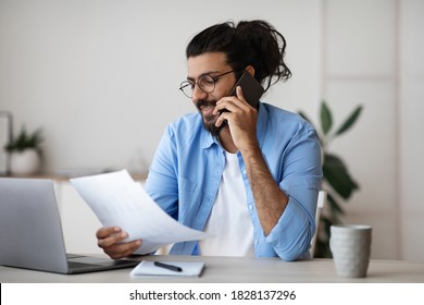 Handsome Indian Freelancer Guy Talking On Cellphone And Working With Papers At Home Office, Discussing Project Budget With Client, Checking Documents And Financial Reports, Sitting At Desk, Copy Space - Shutterstock ID 1828137296
