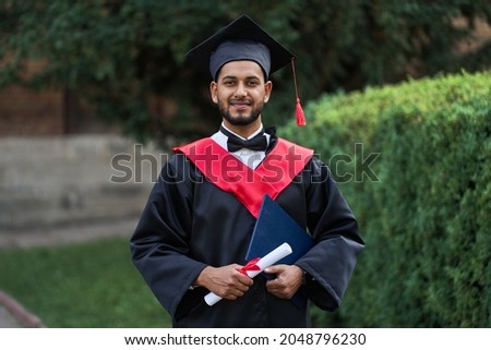 Handsome indian graduate in graduation glow with diploma looking at camera.