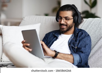 Handsome Indian Guy In Wireless Headphones Using Digital Tablet At Home, Browsing Internet Or Shopping Online, Enjoying Listening His Favorite Music, Relaxing On Couch In Living Room, Closeup