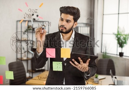 Handsome indian man in formal suit holding digital tablet while writing brilliant ideas on glass wall at corporate office. Concept of business, people and modern gadgets.