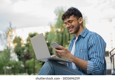 Handsome Indian man using laptop computer, mobile phone, working freelance project online, sitting outdoors. Successful business. Asian student studying, learning language, online education concept