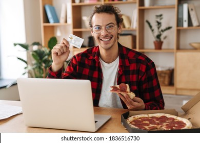 Handsome Joyful Guy Holding Pizza While Eating Pizza And Working With Laptop At Office