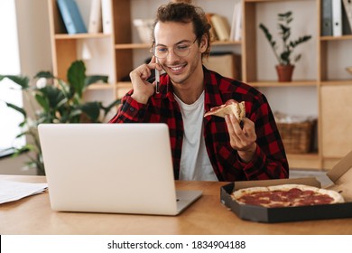 Handsome Joyful Guy Talking On Cellphone While Eating Pizza And Working With Laptop At Office