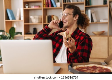 Handsome Joyful Guy Talking On Cellphone While Eating Pizza And Working With Laptop At Office