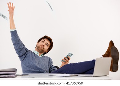Handsome joyful young businessman sitting at the office desk with feet on the desk throwing money over his head. Looking away. Isolated on white background.