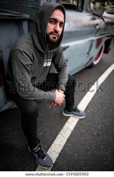Handsome, latin man with dark eyes and black\
hair is based sits on retro, unusual, unique car. Mysterious posing\
model in sport style clothes on the\
road