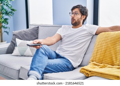 Handsome Latin Man Holding Television Remote Control In Shock Face, Looking Skeptical And Sarcastic, Surprised With Open Mouth 