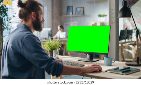 Handsome Long-Haired Bearded Manager Working at a Desk in Creative Office, Using Desktop Computer with Green Screen Mock Up Display. Colleagues Working in the Background in Marketing Agency. - Powered by Shutterstock