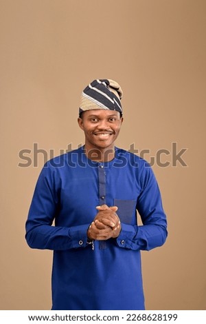 A handsome looking young man pose with hands held to his front looking at the camera, African man wearing blue attires and a cap on isolated background