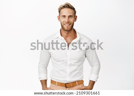 Handsome macho man with blond hair and beard, smiling and looking confident at camera, wearing white shirt, standing against studio background
