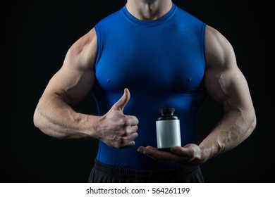 Handsome Macho Man Or Sexy Muscular Guy With Athlete Body And Torso In Blue Sport Vest Holds Pill Jar For Drud, Steroid, Vitamin Or Doping Shows Thumb Up On Black Background