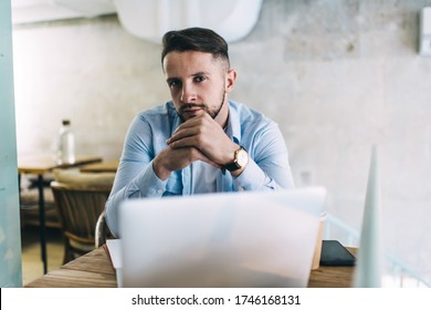 Handsome male with beard and brooding face sitting at table with laptop mobile phone and cup of hot drink in cafeteria - Shutterstock ID 1746168131