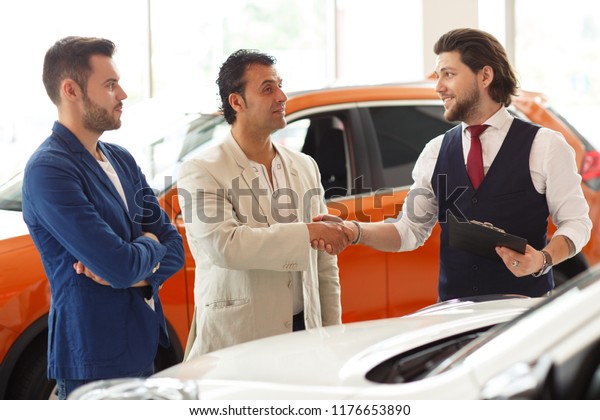 Handsome male client, standing next to\
brand-new car with friend and shaking hand to professional salesman\
at dealership purchasing new car. Two men looking at each other\
happy with\
cooperation.