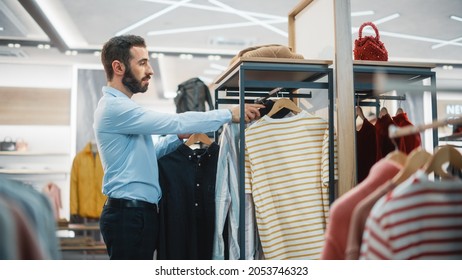 Handsome Male Clothing Store Assistant Works In Fashionable Shopping Mall. Professional Shop Sales Retail Assistant Hangs New Colorful Collection With Sustainable Casual Design On Display Racks.