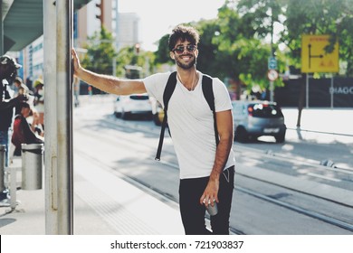 Handsome male with dark curly hair wearing sunglasses is standing on the street with a cup of coffee on a sunny day. Bearded guy wearing casual clothes enjoys relaxing on a summer weekend.