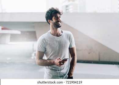 Handsome Male With Dark Curly Hair Wearing Casual Clothes Is Standing With A Mobile Phone In His Hand On Blurred Urban Background. Model Look Man Is Talking On A Smartphone Via Earphones.