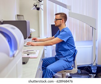 Handsome Male Dentis Using Computer In A Room With Medical Equipment On Background.