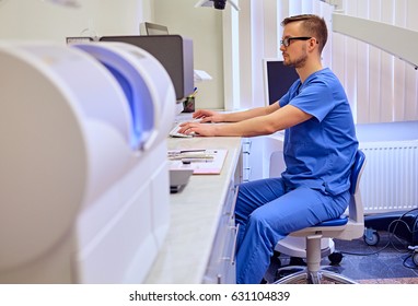 Handsome Male Dentis Using Computer In A Room With Medical Equipment On Background.