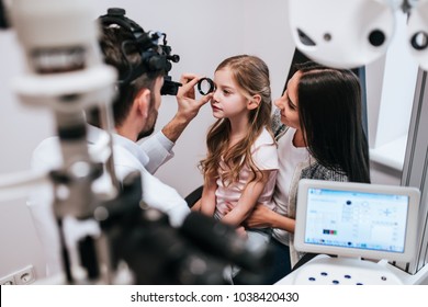 Handsome male doctor  ophthalmologist is checking the eye vision of little cute girl in modern clinic. Mom with daughter in ophthalmology clinic.