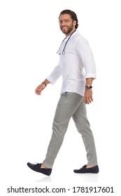 Handsome Male Doctor Is Walking, Looking At Camera And Smiling. Side View. Full Length Studio Shot Isolated On White.