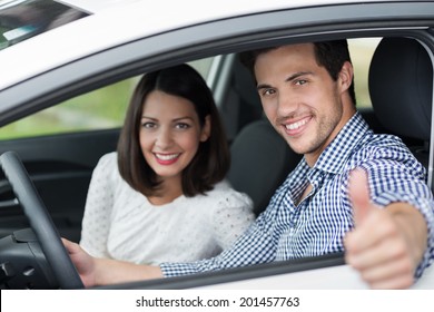Handsome male driver giving a thumbs up through the car window as he drives along with his beautiful young wife or girlfriend as a passenger
