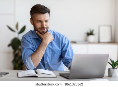 Handsome Male Entrepreneur Using Laptop, Sitting At Desk In Home Office, Looking At Computer Screen With Pleased Face Expression, Having Business Ideas, Enjoying Working Remotely, Selective Focus