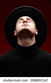 Handsome male fashion model in stylish black sweater and hat poses with closed eyes and his head tilted back on a red studio background. Men's fashion and style. 