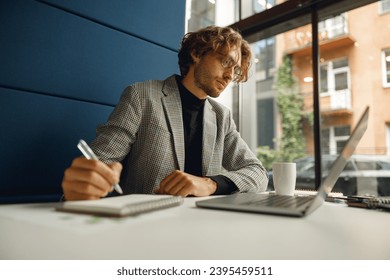 Handsome male freelancer making notes during working on laptop sitiing desk in coworking - Shutterstock ID 2395459511