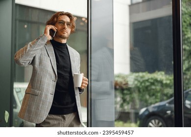 Handsome male freelancer is talking phone and drink coffee standing in modern coworking space - Shutterstock ID 2395459457