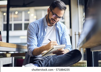 Handsome Male Freelancer Writer Creating Advertising Article For Marketing Company Noting Best Ideas Into Diary Before Finishing Publication Fro Release Working Hard In Morning Sitting In Cafe