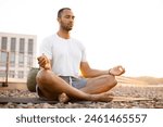 Handsome male meditating in lotus position while sitting on mat on roof of modern house. Young athletic man doing yoga at sunset on urban background.